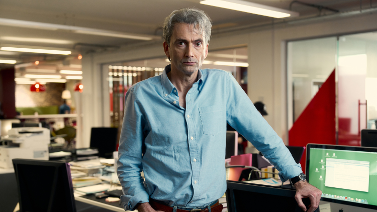 David Tennant, a white man in his fifties, looks into the camera, unsmiling, wearing a blue shirt, stood in what appears to be a newsroom