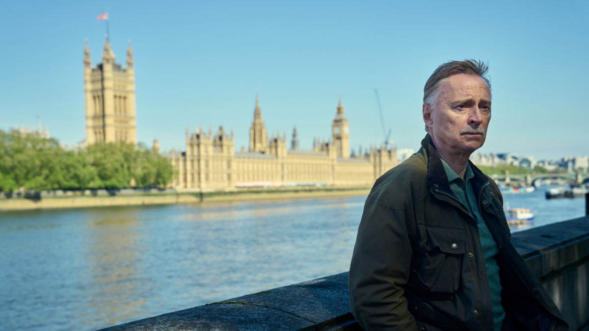 Robert Carlyle, a white man in his sixties, stands by the River Thames, back turned to the Houses of Parliament, out of focus and on the other side of the river