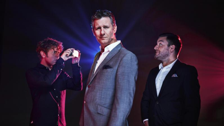 Comedians Josh Widdicombe and Adam Hills stand in line with presenter Alex Brooker in front of blue and red lights