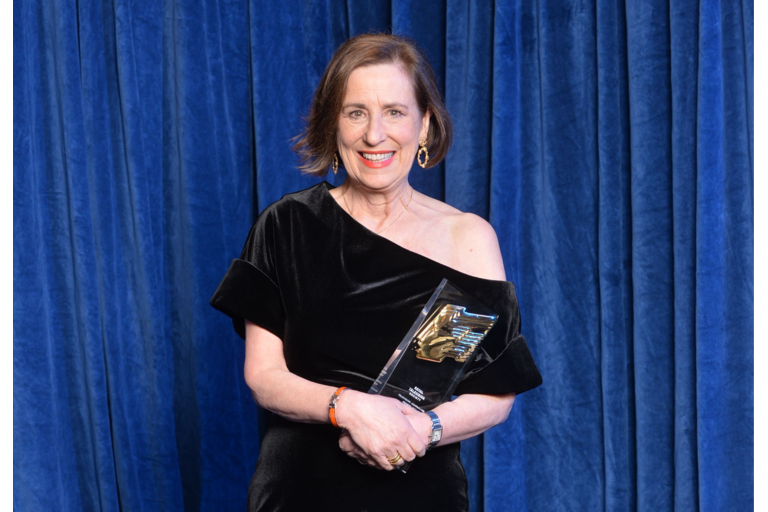 Kirsty Wark, a woman in her late 60s, has brown bobbed hair and is  wearing a velvet off the shoulder gown. She holds her award in front of a blue curtain.