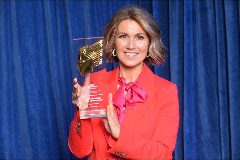 Susanna Reid wears a red suit with a pink bow tied around her neck. She has brown bobbed hair with blonde highlights. She holds an award in front of a blue curtain.
