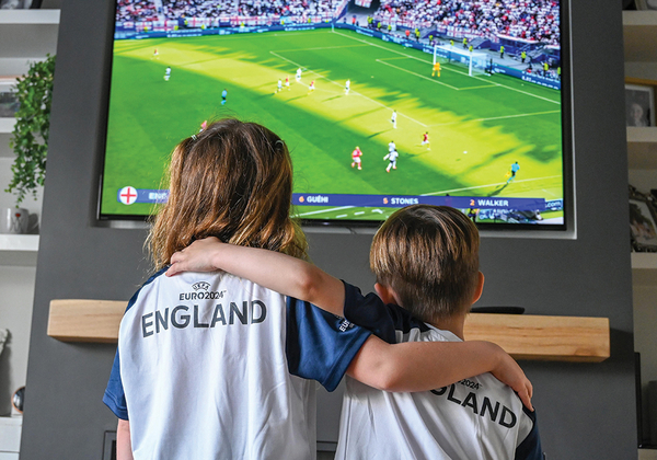 Two kids wearing England football shirts watch England play in Euro 2024 on TV 