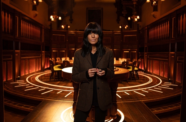 Claudia Winkleman, a woman with a light skin tone, and dark hair with a long fringe, stands in front of a large round table surrounded by seats