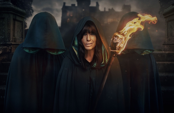 Claudia Winkleman stands in the middle with a green cloak holding a fire on a stick, with two figures either side of her, covered by their hoods