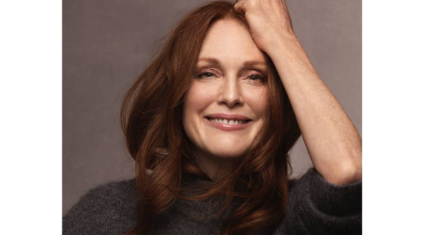 Julianne Moore with auburn hair in a grey jumper stands in front of a brown screen