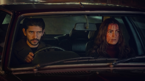 Ben Whishaw and Keira Knightley, both in their late 30s/early 40s, sit side by side in a car. Both have light skin tones, Ben has dark hair and a beard. Keira has long light brown hair that's waved, and she is covered in blood.