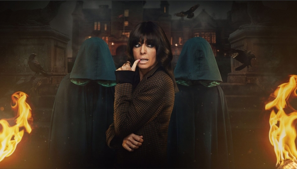 Claudia Winkleman stands in front of two hooded figures in front of a castle at night. Flames are burning on the lower left and right corners