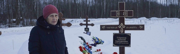 An elderly woman stands in front of three graves, which have writing in Cyrillic 