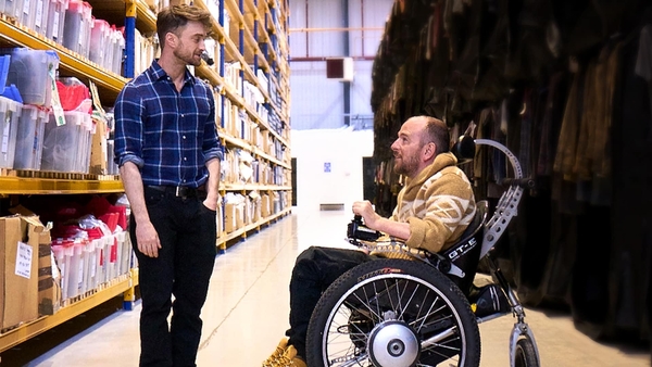 Daniel Radcliffe (left) talks to David Holmes (right) talk in a warehouse, with David in a wheelchair