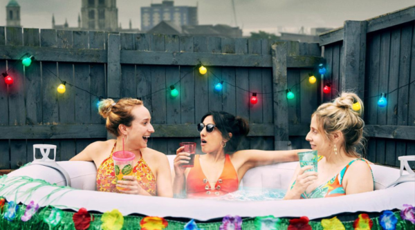 In a still from sitcom Hullraisers, three women sit in a hot tub, talking while drinking from brightly coloured glasses.
