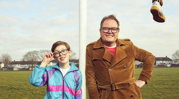Alan Carr stands to the right of a goal post, with his younger self to the left. Someone is sitting on the post, but the only part of them in the photo is their right foot, hanging above the older Alan