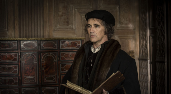 Mark Rylance playing Thomas Cromwell in Tudor dress stands in front of ornate wooden panels