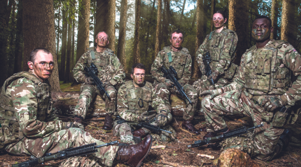 A group of soliders in camouflage dress and war paint sit and stand in a forest