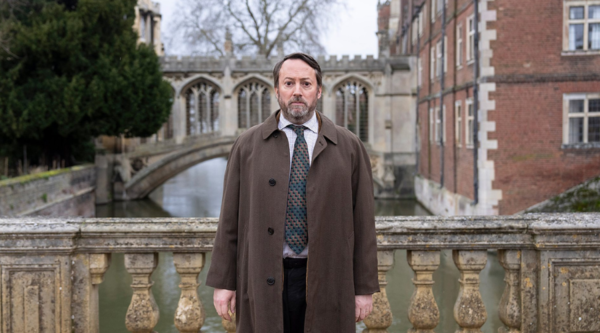 David Mitchell, in character as John ‘Ludwig’ Taylor, stands on a bridge in Cambridge, behind which is the famous Bridge of Sighs