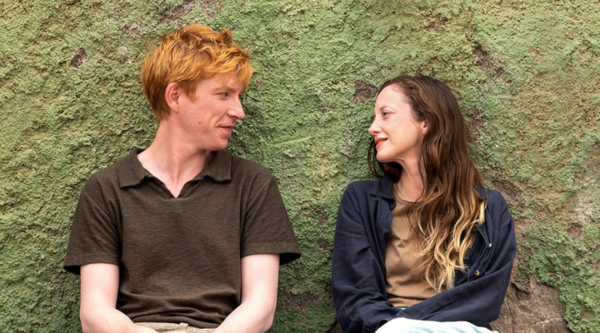 Domhnall Gleeson and Andrea Riseborough sit looking at each other in front of a stone, moss green-coloured wall
