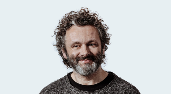 Michael Sheen looks into the camera, smiling, in front of a pale bluish grey background