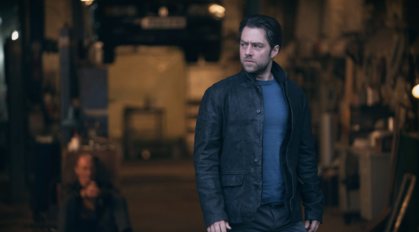 A man stands outside an open building, perhaps a garage, in which a man sits, out of focus