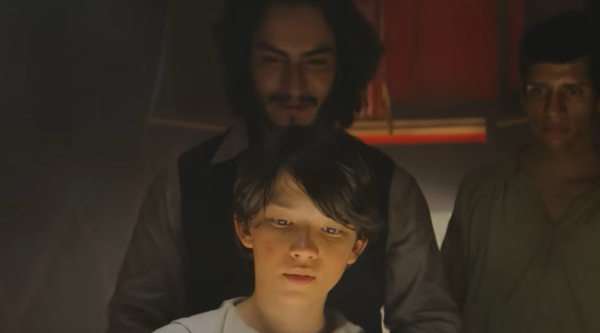 A man stands behind a boy, as something shiny off-camera illuminates his face