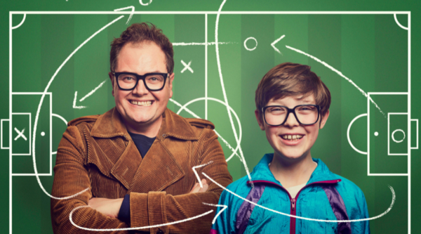 Alan Carr and Young Alan (Oliver Savell) look into the camera, smiling, in front of a graphic of a football field, complete with arrows and annotations