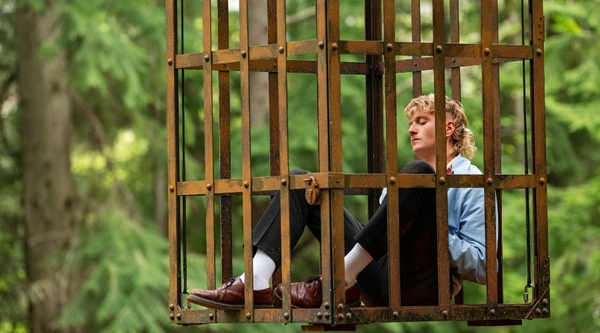 A white man with blonde hair sits down in a hanging cage, looking dejected