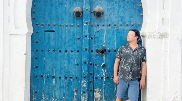 Jamie Oliver is stood against a blue weathered door that is twice his height and curved at the top