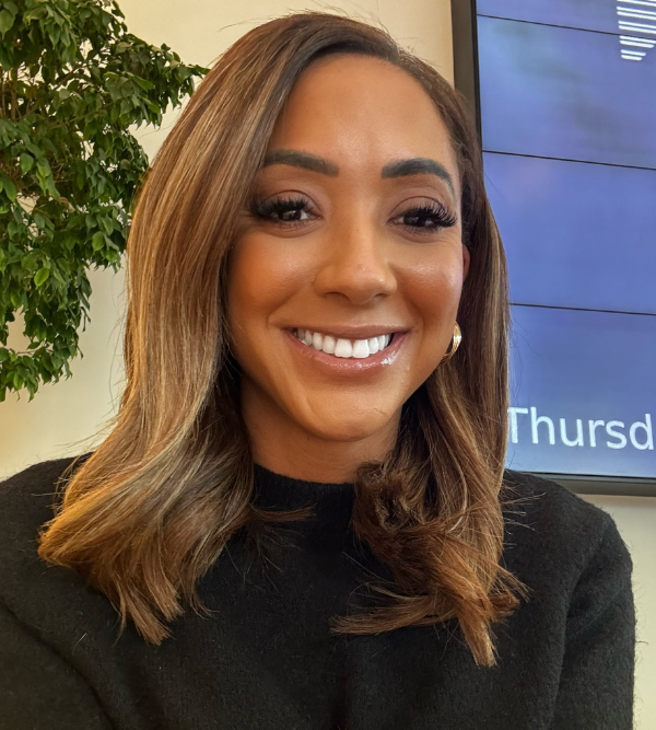 A woman with shoulder-length, light brown hair is smiling. She is wearing a black top and gold hoop earrings. The background includes a partial view of a large screen and a green potted plant.