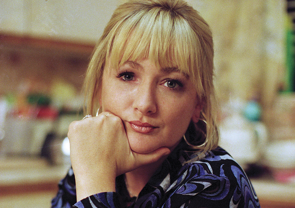 A headshot of Caroline Aherne, a white woman with blonde hair wearing a blue and black patterned shirt