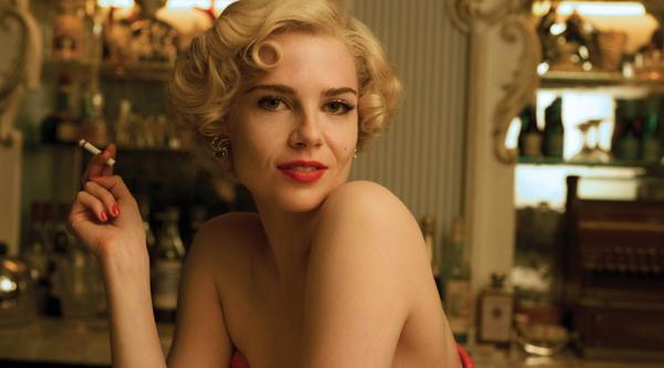 A white woman in her 30s with blonde hair wears a red dress and smokes a cigarette in an old pub