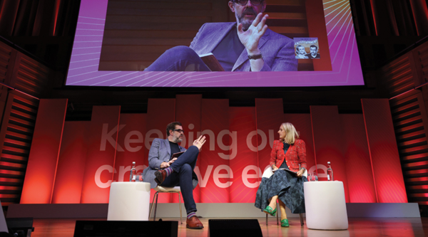 Richard Osman and Marina Hyde talk onstage at the RTS London Convention, beneath a large screen displaying a live feed of Osman