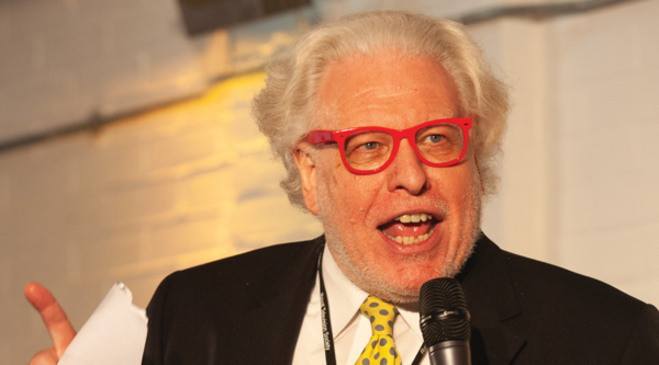 Simon Albury speaking publicly, wearing distinctive red glasses