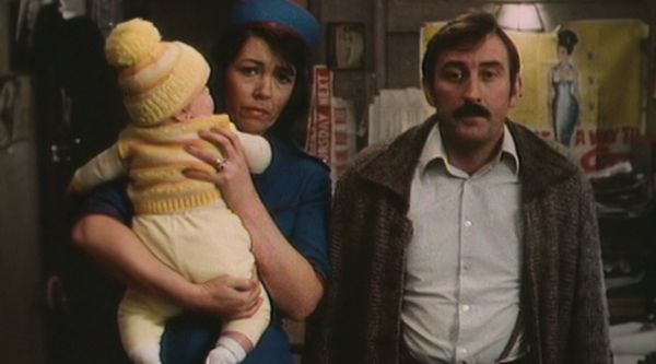 Iola Gregory and Dafydd Hywel stand looking into the camera, the former holding a baby