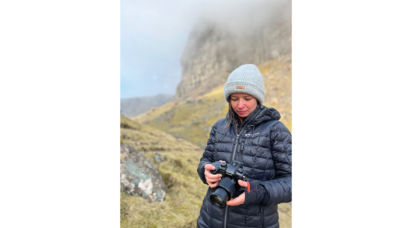 Ellie Green holds a camera while standing on a grassy and rocky mountain