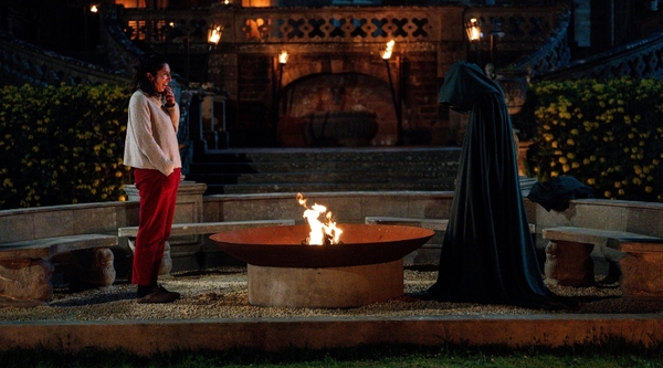 A white woman with dark hair stands by a firepit outside a castle and opposite a fully cloaked person