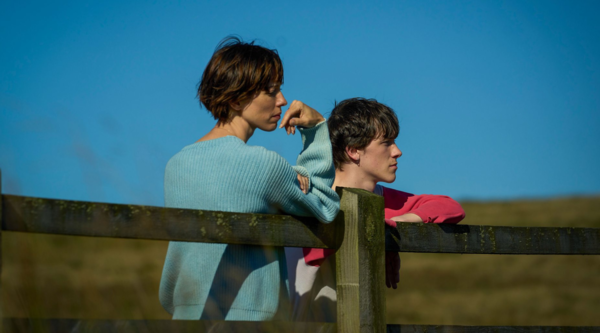  Rebecca Hall and Ollie West stand against a fence in a field, the sky behind them is blue and cloudless. Hall is in her early 40s, with short brown hair, light to medium skin tone and a turquoise jumper, Ollie West is in his late teens