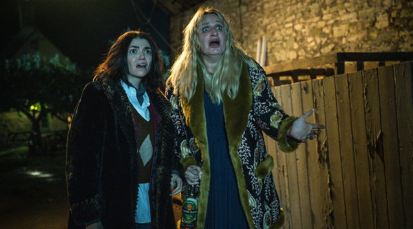 Selin Hizli and Daisy May Cooper stand outdoors at night, looking surprised and slightly horrified at something off-camera