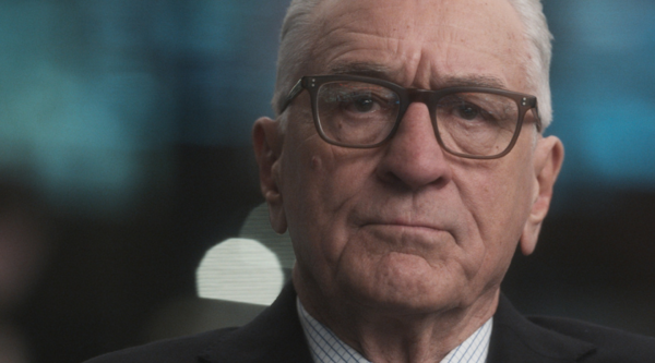 Robert De Niro, a white man in his eighties, is unsmiling in glasses and a suit, a blue and dark backdrop blurred behind him