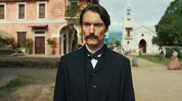 Aureliano looks stern dressed in a formal suit in the fictional town of Macondo, with a Church pictured behind him