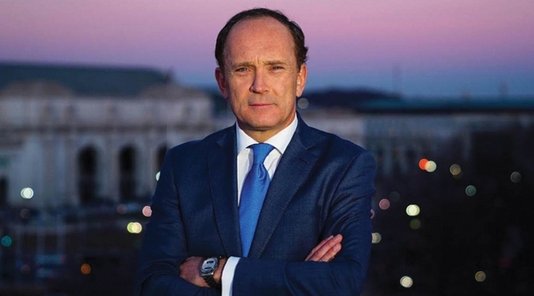 A white man with dark hair wears a navy suit and stands with his arms crossed against a purple sunset backdrop