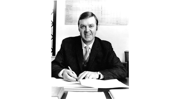 A black and white photo of a white man in a suit sitting at a desk and writing on a notepad