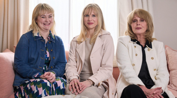 Lucy Punch, Joanna Lumley and Philippa Dunne sit on a sofa, smiling to the camera. Lumley and Punch wear light-toned, expensive-looking clothing, whereas Dunne wears a denim jacket and dark multicoloured dress