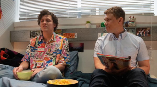 Dylan Llewellyn and Jon Pointing, both white men in their 30s, sit on a bed, with Dylan wearing a name sticker reading "JACK" and a multi-coloured button-up shirt. Jon wears a pale blue button-up shirt