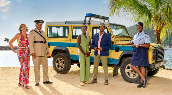 The cast stand in front of a police 4x4 on a tropical beach, a palm tree and verdant hills behind them