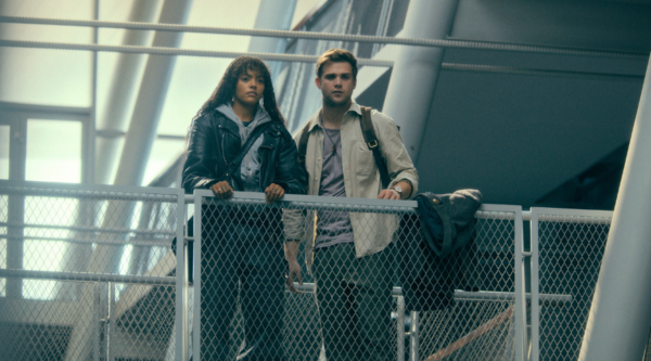 Quintessa Swindell, a biracial woman in her 20s, and Leo Woodall, a white man in his late 20s, stand together on a walkway indoors