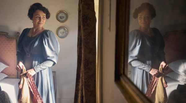 Keeley Hawes, a white woman in her forties, holds a sheet while looking out the window, smiling and wearing a blue 19th century everyday-wear dress