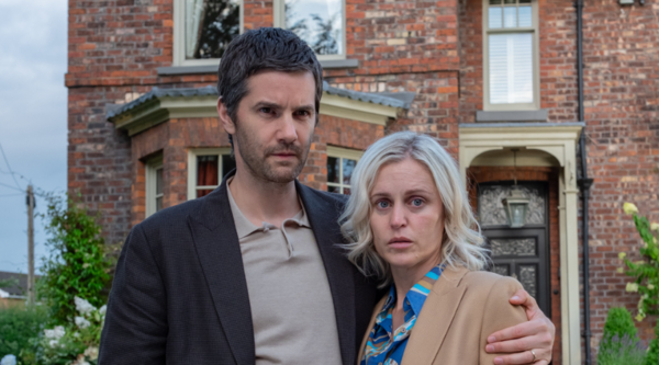 Denise Gough, a white woman in her forties, and Jim Sturgess, a white man in his forties, stand arm in arm outside a large-ish suburban red brick house, unsmiling