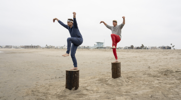 The two stand on wooden plinths on a beach, adopting a crane-like pose