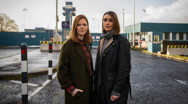 The pair stand together in front of a fortified entrance to a compound of some kind with tall blue fences