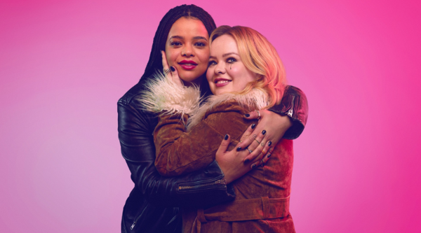 Lydia West, a British-Caribbean woman in her thirties, hugs Nicola Coughlan, a white woman in her thirties, against a pink backdrop. Coughlan's mascara is running slightly, seemingly from crying