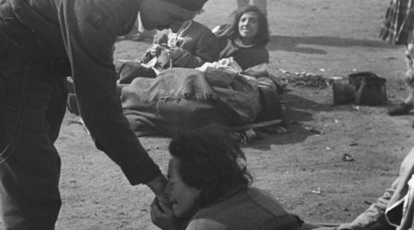 In a grainy black-and-white photo, a man in army uniform extends a hand to a woman who has pressed it against her face and is crying. A woman looks on behind both of them