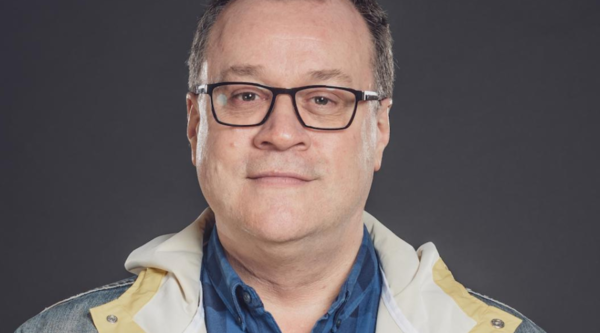 Russell T Davies, a white man in his sixties, looks into the camera, smiling, against a grey backdrop